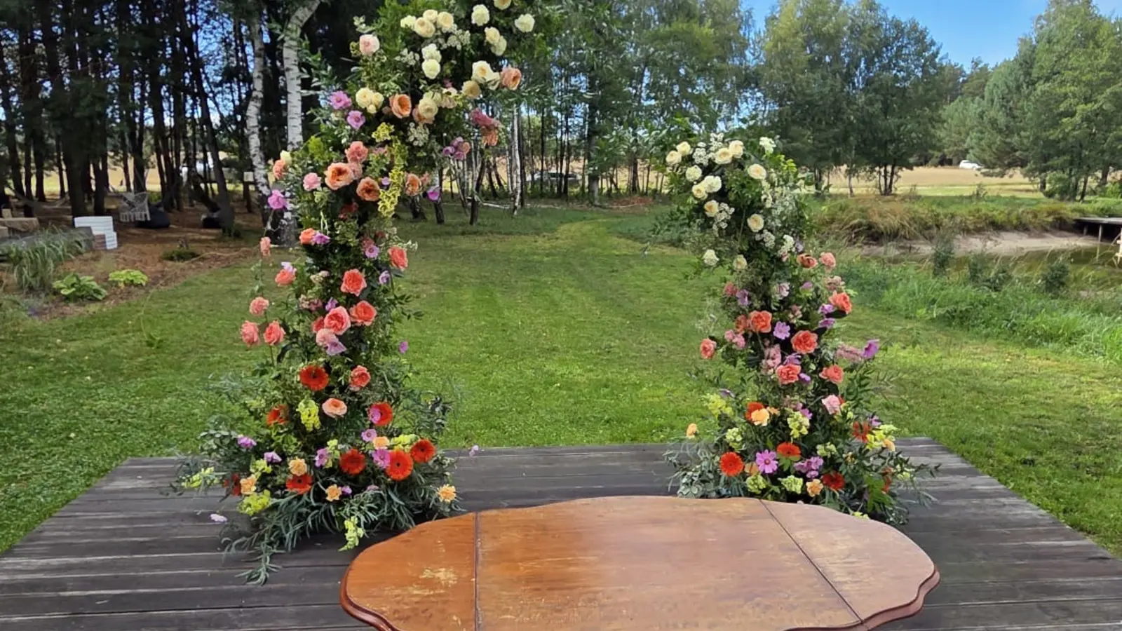 Ceremonia na drewnianym podeście na łące: dwie kolumny kwiatowe z róż i drobnych kwiatów w brzoskwiniowych, różowych i fioletowych kolorach po bokach, przed nimi drewniany stolik, w tle brzozy i sosny.