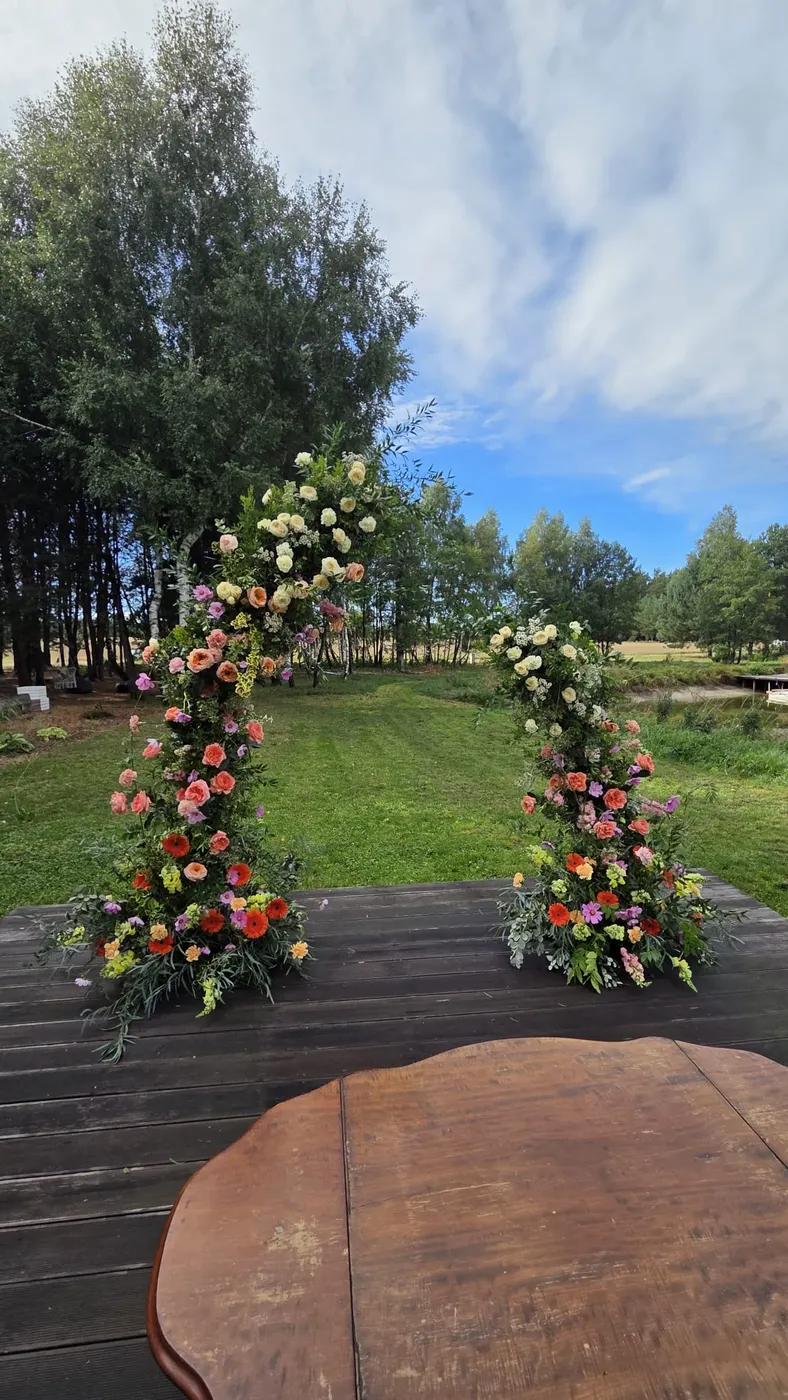 Dwie kolumny kwiatowe w kolorach brzoskwini, czerwieni i bieli stoją na drewnianym podeście w ogrodzie, przed nimi drewniany stolik do ceremonii.